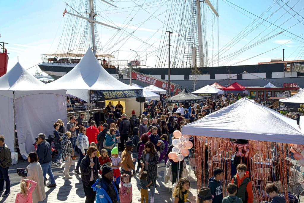 outdoor food festival with tents and people, with a ship in the background