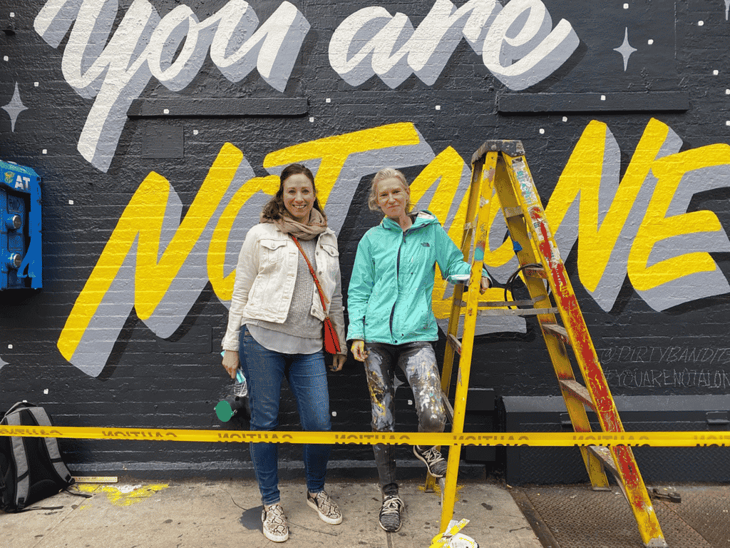 Two women artists in front of "You are not alone" mural