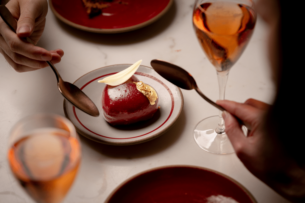 Celebrating Valentine's Day at the Seaport, two people enjoy a red dessert topped with white chocolate and gold leaf, holding spoons. Two glasses of rosé wine are on the table.