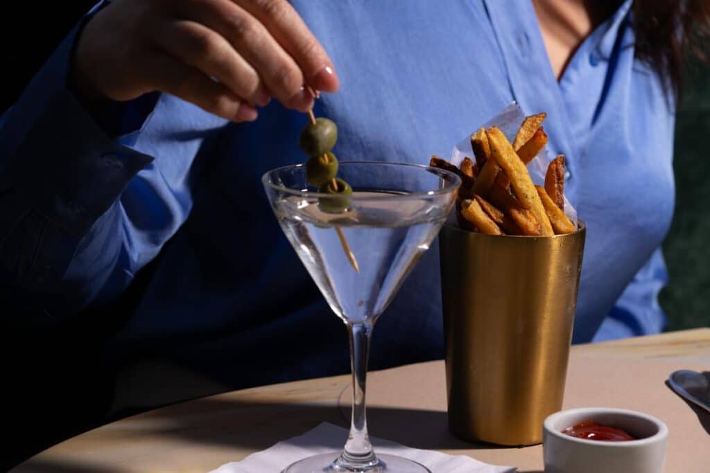 A person in a blue shirt dips olives into a martini glass beside a cup of fries and a small dish of ketchup on a table.