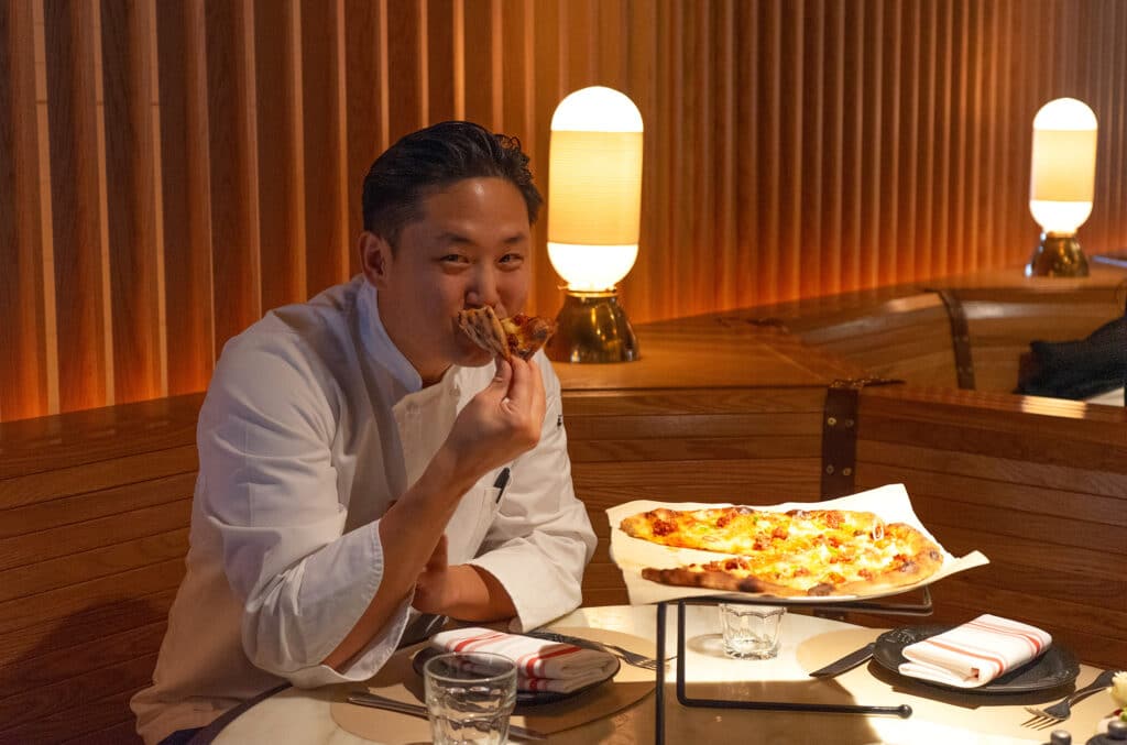 A person in a chef's coat eats a slice of pizza in a restaurant with wooden decor and warm lighting.