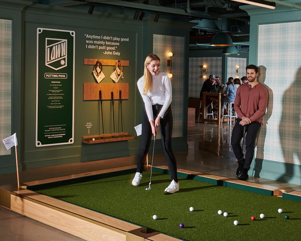 a woman putts on an indoor green at lawn club while a man looks on