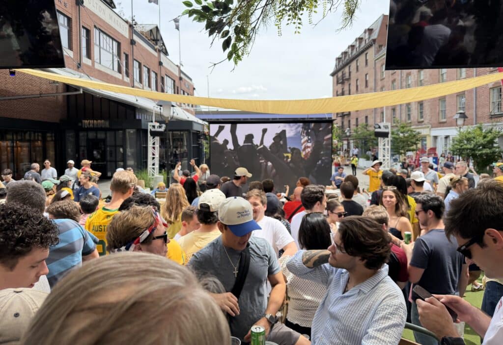 a crowd of people watch a sporting event at sadie's garden bar's 18 foot outdoor screen at the seaport