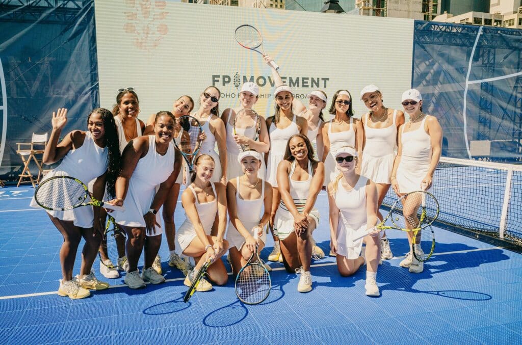 a group of women stand on a blue tennis court, all wearing white, and holding various tennis racquets and balls.