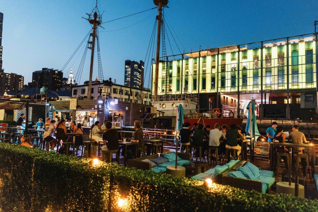 Cobblefish, an outdoor bar and restaurant on pier 16, with a view of the illuminated Pier 17 building and NYC skyline behind it.