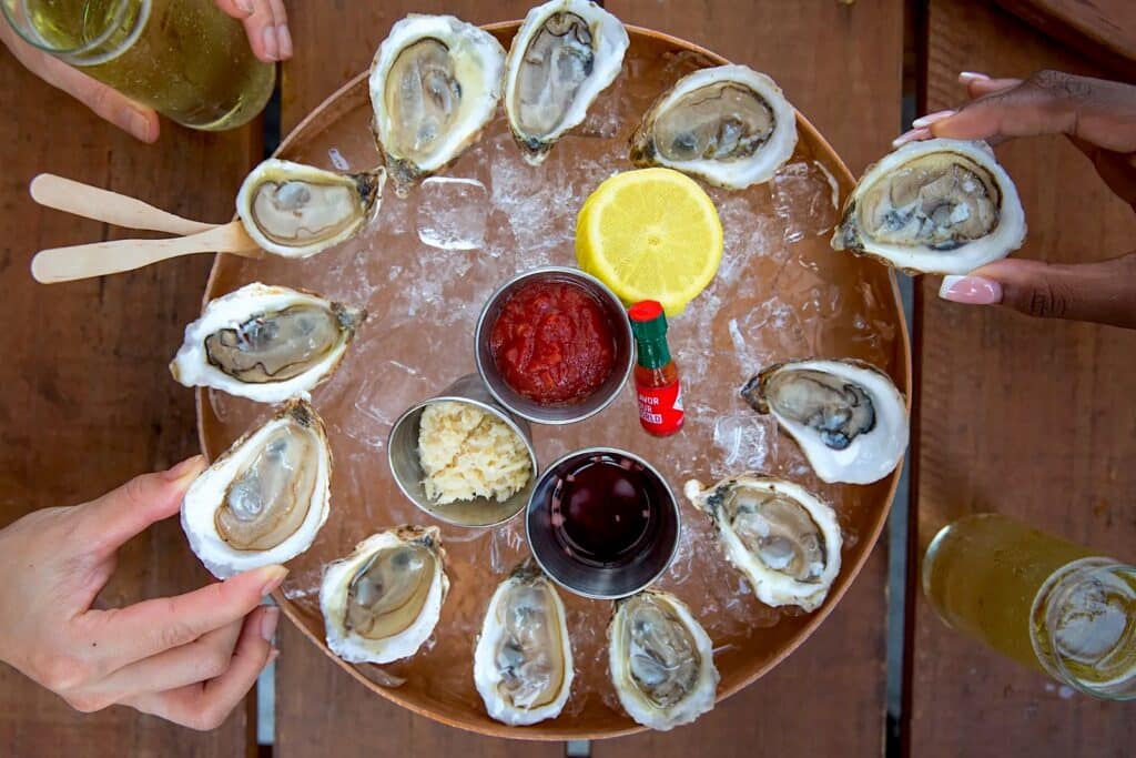 a plate of oysters on a bed of ice, with accoutrement, including a mini bottle of tabasco. two hands can be seen reaching into frame and grabbing oysters