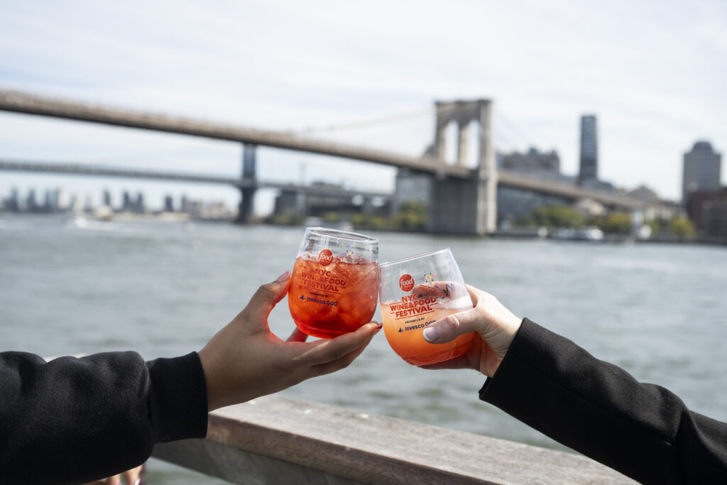 two people clink NYC Food and Wine Festival glasses with cocktails in front of the brooklyn bridge