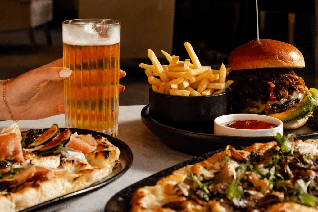 a hand reaches for a glass of beer on a table, surrounded by a burger and various pizzas, as part of urban cove's happy hour menu at the seaport