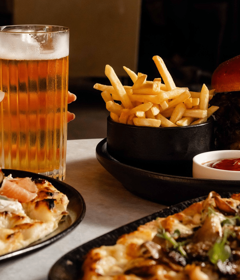 a hand reaches for a glass of beer on a table, surrounded by a burger and various pizzas, as part of urban cove's happy hour menu at the seaport